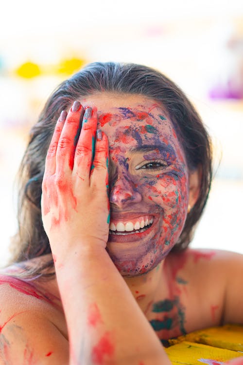 Happy young female with colorful paints on face looking at camera while covering half of face with hand on street against blurred background