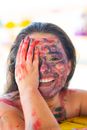 Happy young female with colorful paints on face looking at camera while covering half of face with hand on street against blurred background