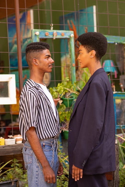 Free Side view of African American couple in stylish clothes looking at each other while standing near metal fence on street Stock Photo