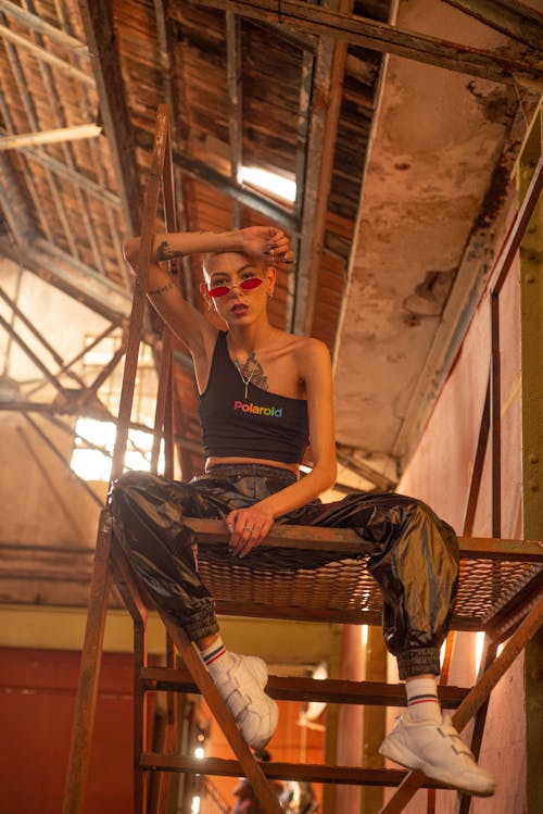 Low angle of young bold female in trendy outfit and sunglasses sitting on metal construction in abandoned building and looking at camera