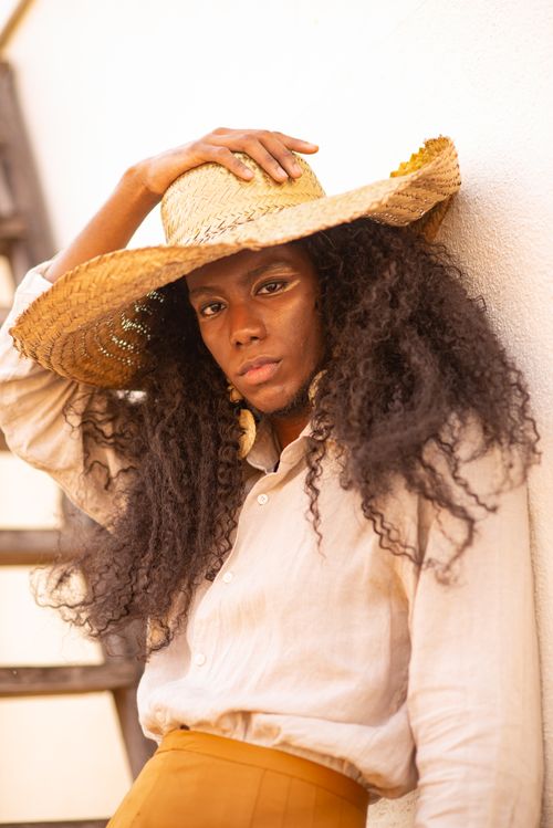 Free Young African American female in trendy outfit and straw hat touching head and looking at camera while leaning on white wall in daytime Stock Photo