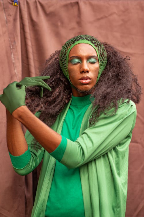 Extravagant African American female with curly hair and closed eyes wearing colorful green clothes gloves and thin net on head touching hair while standing against brown curtain in studio