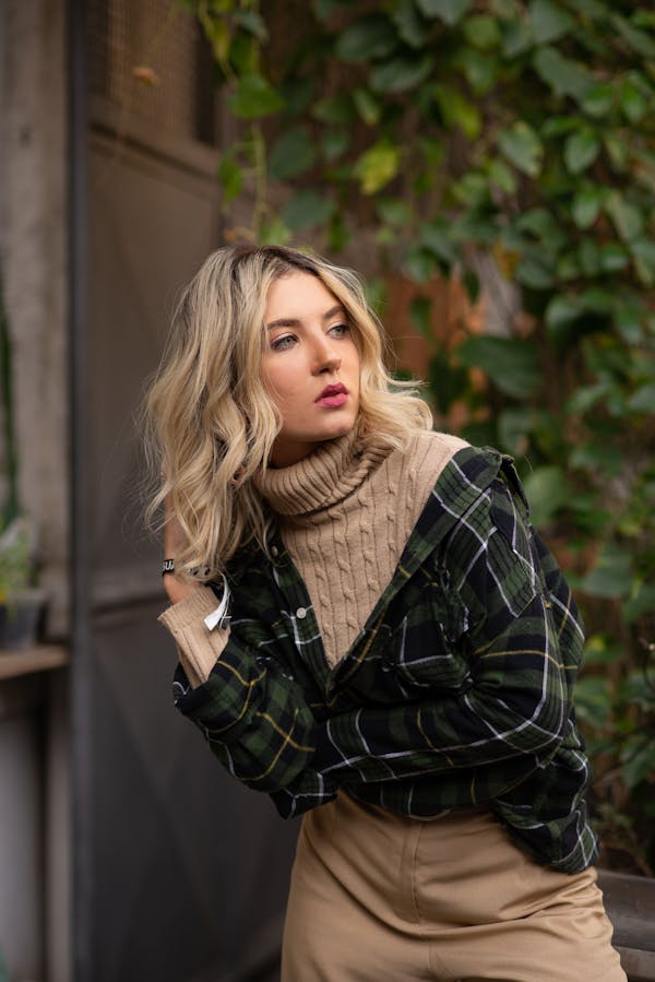 Charming young female with fair hair wearing knitted jumper and checkered shirt looking away while standing on blurred background of building and green climber plant