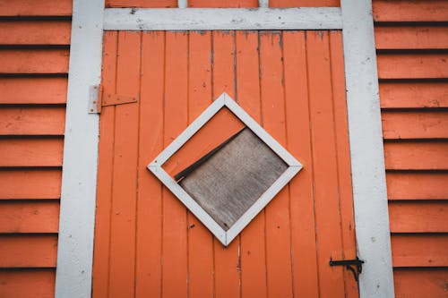 Weathered wooden orange door with rhombus detail and metal hinge of shabby building with boards located on street in town