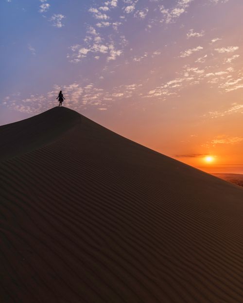 Free A lone figure stands atop a sand dune in Huacachina as the sun sets, creating a dramatic silhouette. Stock Photo