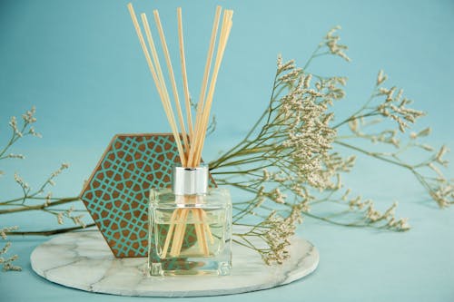 Aroma sticks in glass diffuser bottle with decorative Gypsophila branch placed on marble stand near geometric decor on blue background