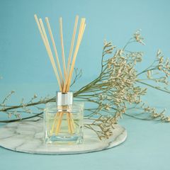 A square glass reed diffuser with natural reeds on a marble stand against teal