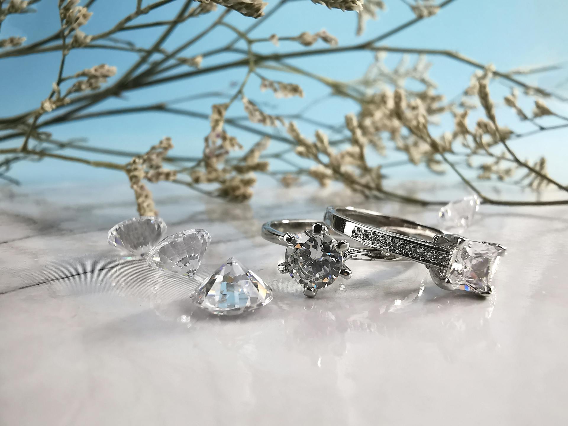 Elegant silver wedding rings with diamonds and crystals placed on shiny white table with twigs of tree on light blue background