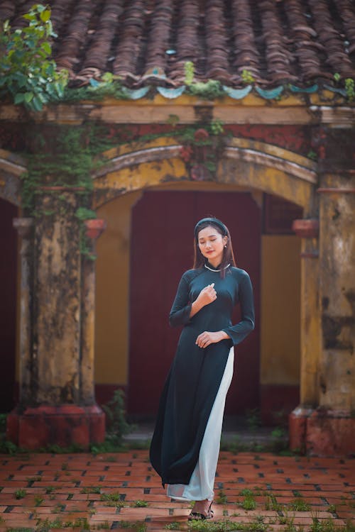 Free Full body of Asian female in traditional dark dress standing near ethnic building with closed eyes in sunny day Stock Photo