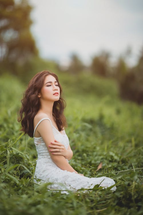 Dreamy long haired Asian lady in white dress sitting in grassy lawn and closing eyes