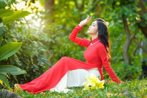 Side view full body of young ethnic female with long black hair in red summer dress sitting on green lawn with arm raised