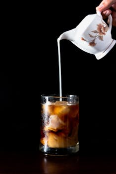 A hand pours cream from a floral pitcher into an iced coffee against a dark background.
