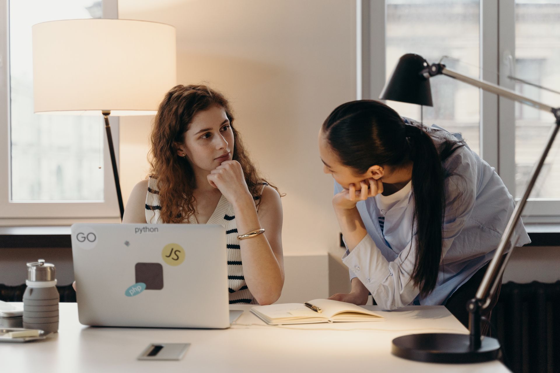 Two businesswomen collaborating on a project with a laptop and notebook in an office setting.