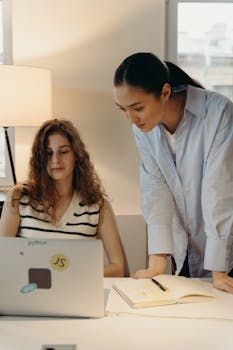 Two women collaborating on a project in a modern office environment with a laptop and notebook.
