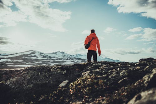 Free A Man in Orange Jacket Standing Near the Snow Covered Mountains Stock Photo