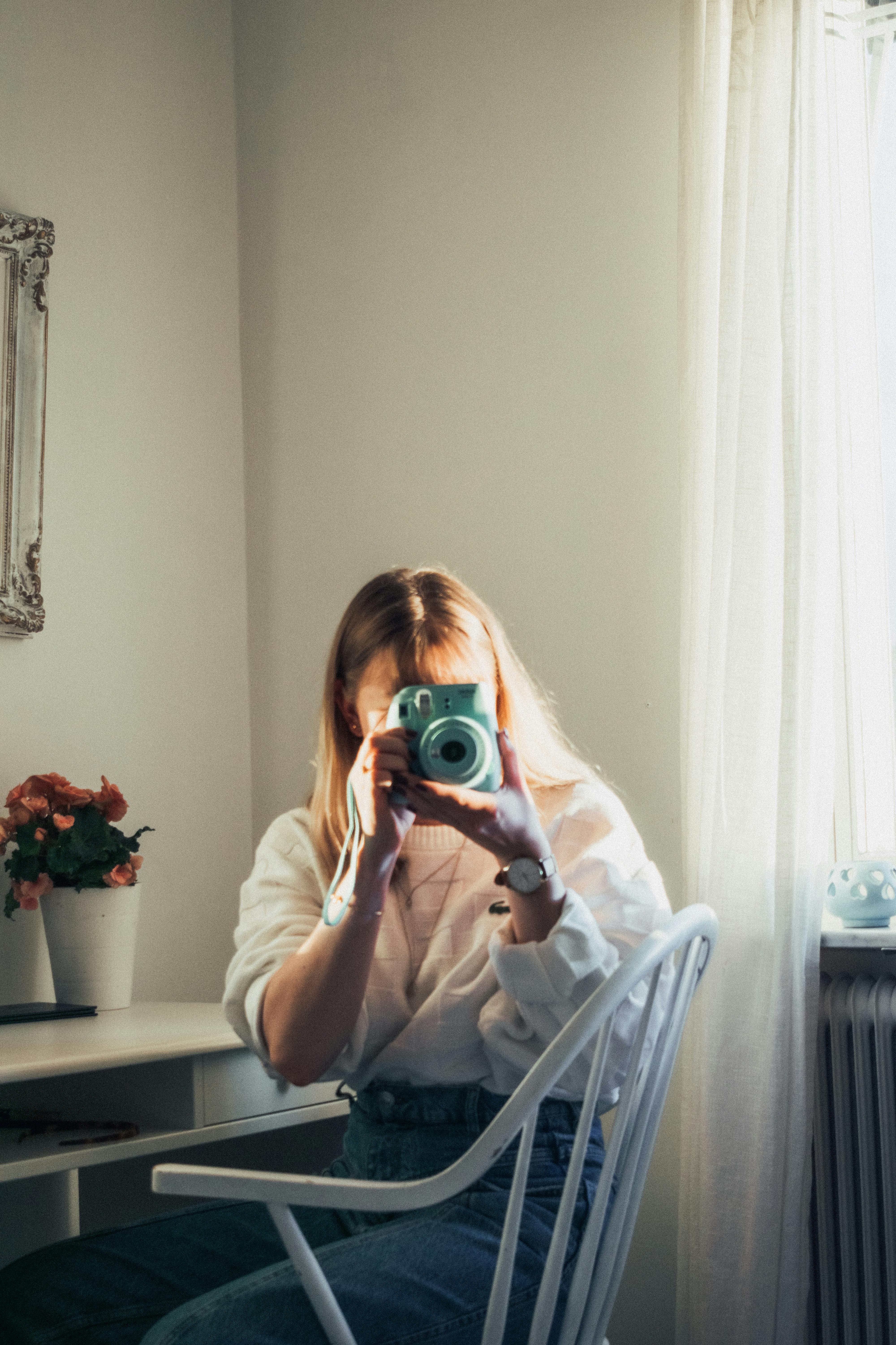 A woman with a watch takes a photo in a sunlit room with flowers and a mirror.