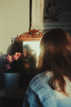 A woman in a blue sweater reflects in a mirror with sunrays illuminating her hair and pink flowers beside her.