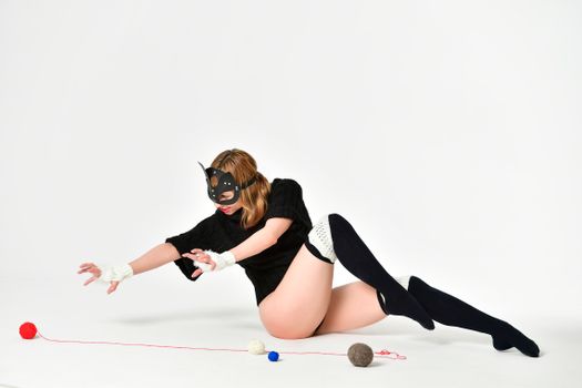 Full body of female in catwoman costume sitting on floor with colorful balls of thread on white background in studio