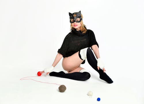 Full body of sensual female in catwoman outfit with mask looking at camera while sitting on white background with colorful balls of thread