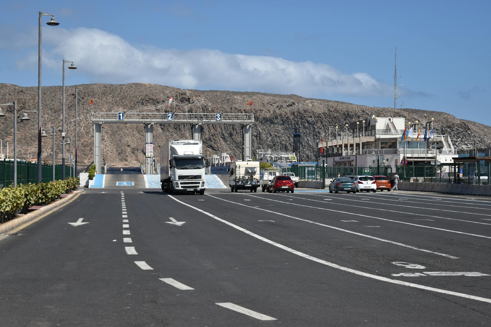 A ferry terminal with trucks and cars under a clear blue sky and dry hills in the background.