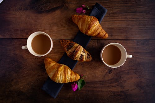Free Three Croissants and Two Cups of Coffee on Wooden Table with Pink Flowers Stock Photo