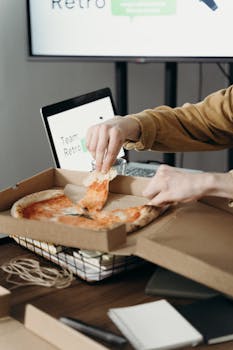 A person enjoys a slice of pizza in an office setting, with a laptop and workspace in the background.