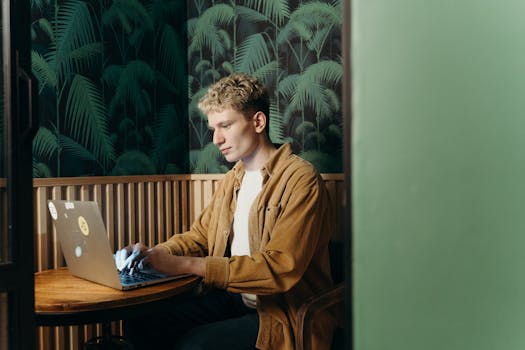 A focused man typing on a laptop in a modern cafe with tropical decor.