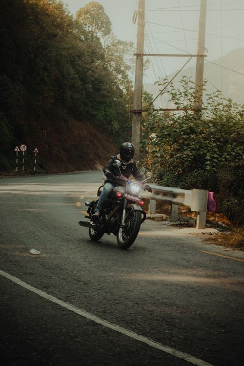 Free A biker in a black jacket rides a motorcycle on a winding road through a forested area, captured during the day. Stock Photo