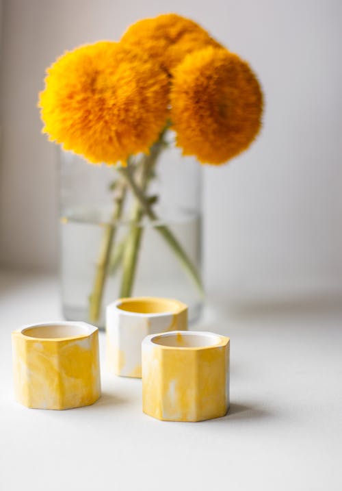 Bunch of vibrant yellow craspedum flowers in glass vase placed on windowsill near decorative candleholders in daylight