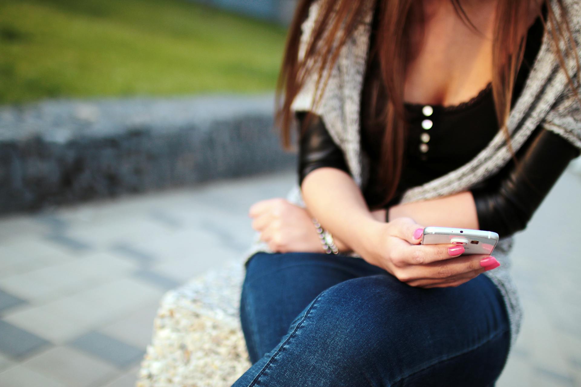 A young woman sitting outdoors using a smartphone, focused on her mobile device.