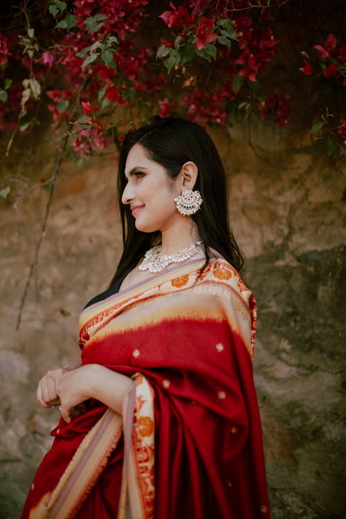 Free Side view of young ethnic female with dark long hair with gold earrings and necklace wearing red traditional Indian sari standing near rocky wall with blooming flowers Stock Photo