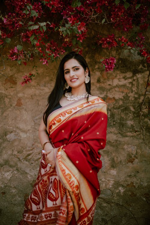 Free Positive young ethnic female with dark long hair in necklace and earrings wearing traditional Indian sari with ornament near stone wall with blooming flowers of bush Stock Photo