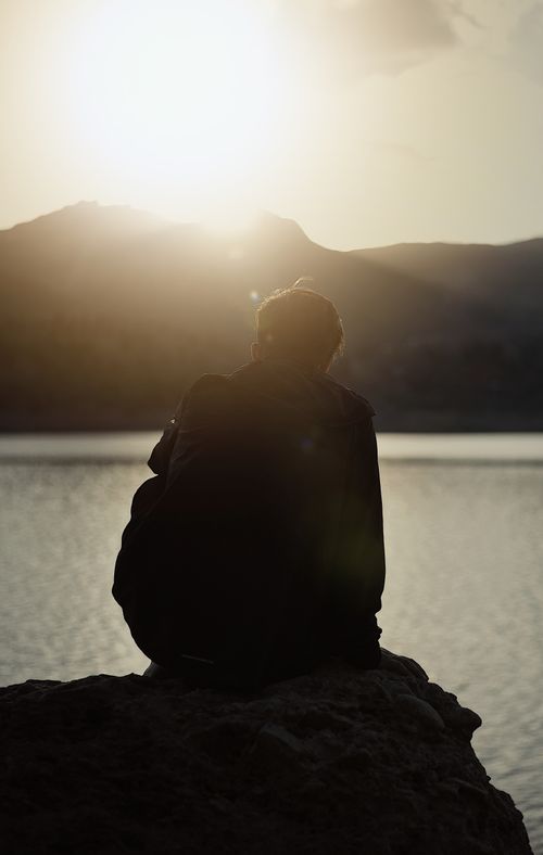 Free A tranquil moment captured as a person sits by the lake, gazing at the sunset over the mountains. Stock Photo