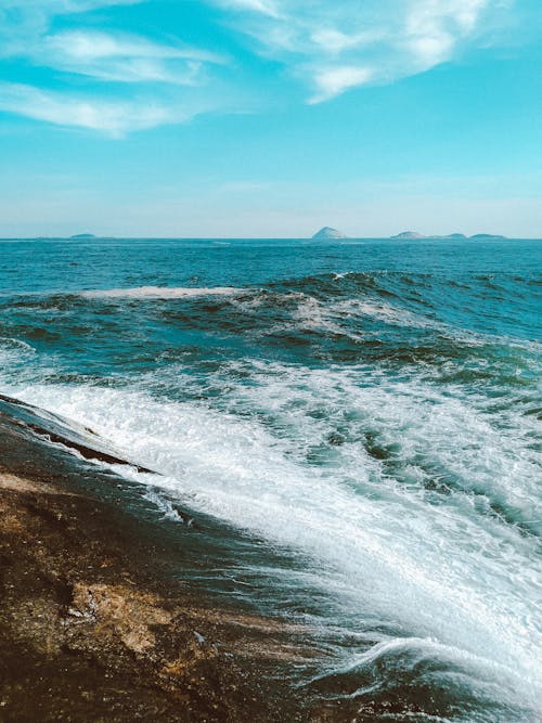 Picturesque view of foamy waves of turquoise ocean crashing on rocky coast in sunny day