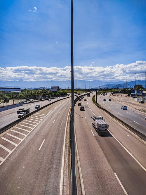 Curvy asphalt roadway with marking lines and vehicles leading to hills under blue sky with white clouds in sunny day