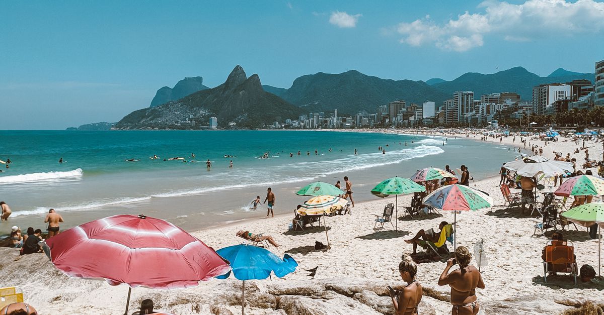 Anonymous tourists sunbathing on sandy shore with parasols against sea and mount under cloudy blue sky in sunlight