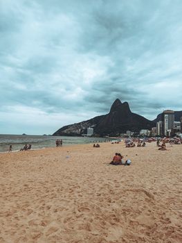 Anonymous tourists on sandy coast against mountain and endless ocean under cloudy sky in city