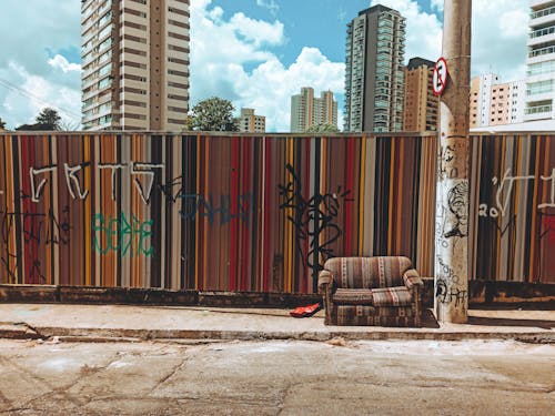 Couch on walkway against fence with letters and contemporary multistory house facades under cloudy sky in sunny city