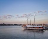 Ships with masts moored on river against city building in harbor under cloudy sky in evening