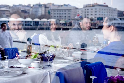 Through glass of smiling friends sitting at served table with dishes and drinks and having conversation in restaurant on coast of sea