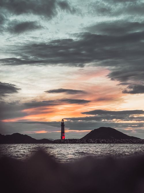 Free A dramatic silhouette of a lighthouse against a colorful sunset sky, reflecting on the sea. Stock Photo