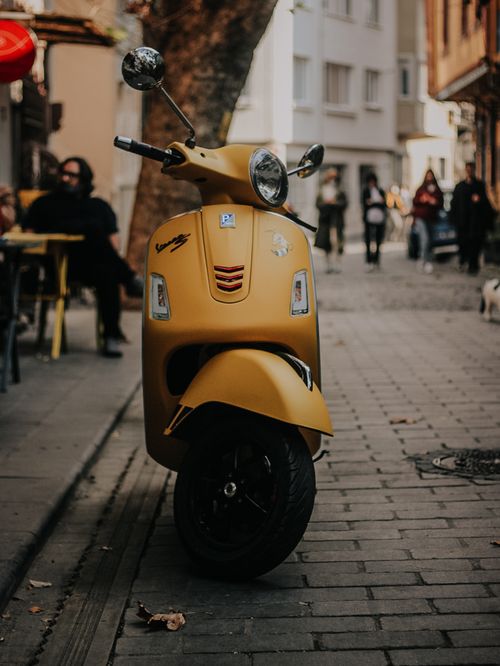 Free A yellow vintage scooter parked on a cobbled urban street with blurred pedestrians in the background. Stock Photo