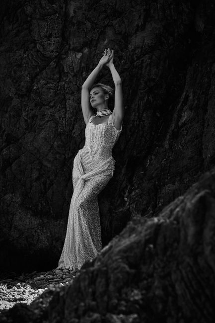 Stunning black and white photo of a model in a glamorous gown posing against a rocky backdrop.