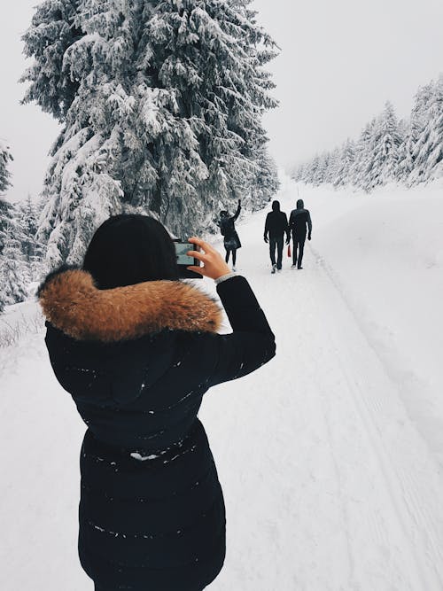 Женщина в черном пальто фотографирует трех человек перед собой во время прогулки по снежному полю