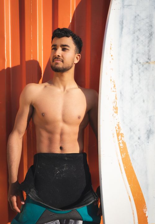 Free Shirtless young man leans on surfboard against vibrant orange wall, exuding confidence. Stock Photo