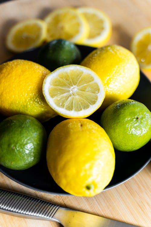 From above of whole and sliced yellow lemons and green limes on plate near knife on wooden table