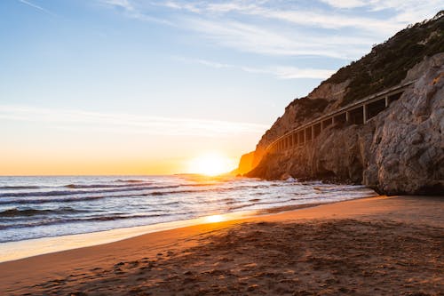 Amazing scenery of waving ocean washing sandy beach near rocky hill against bright sunset sky