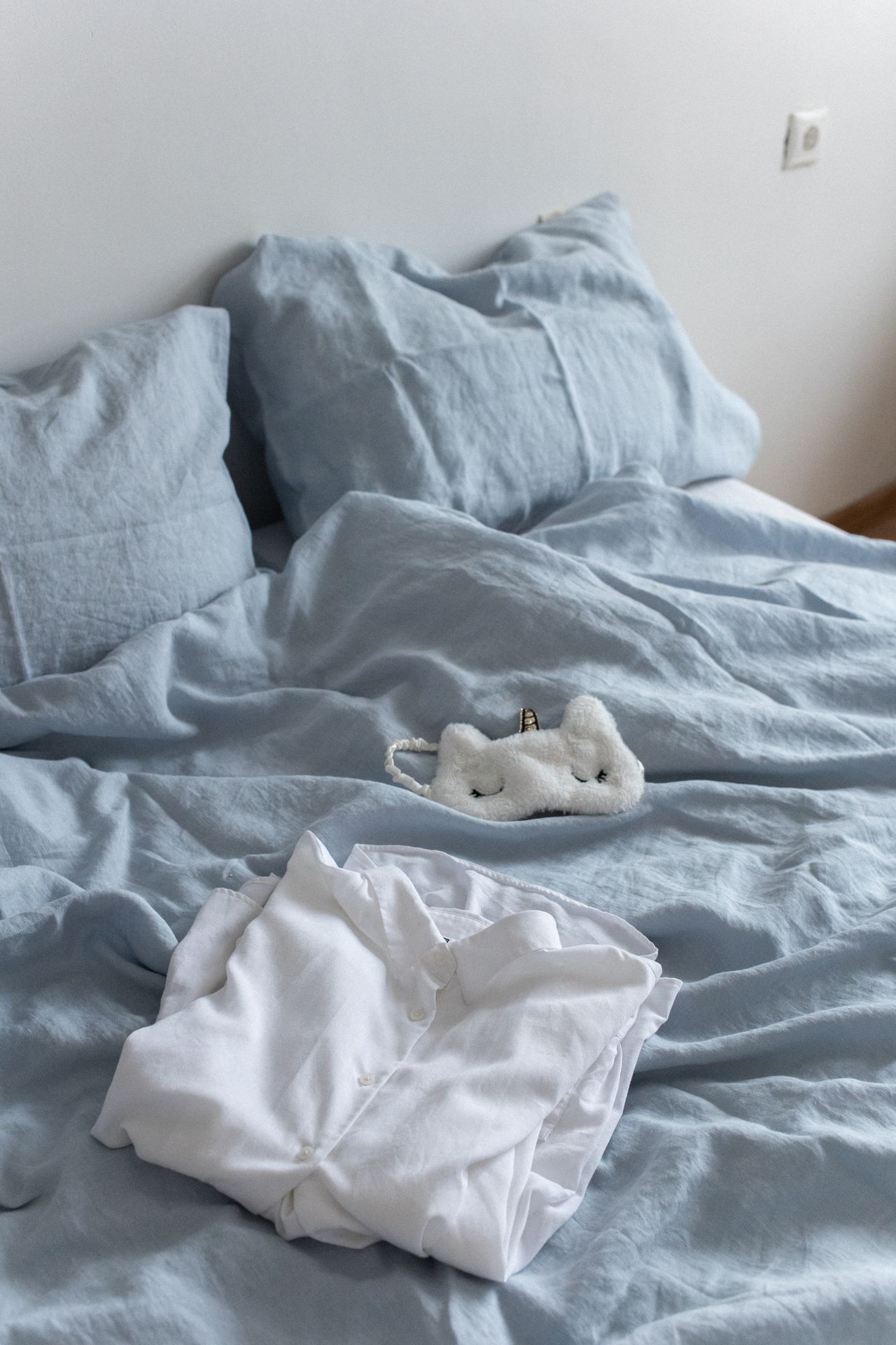Soft light blue linen bedding with rumpled pillows in a calm bedroom with cream walls and warm wood flooring