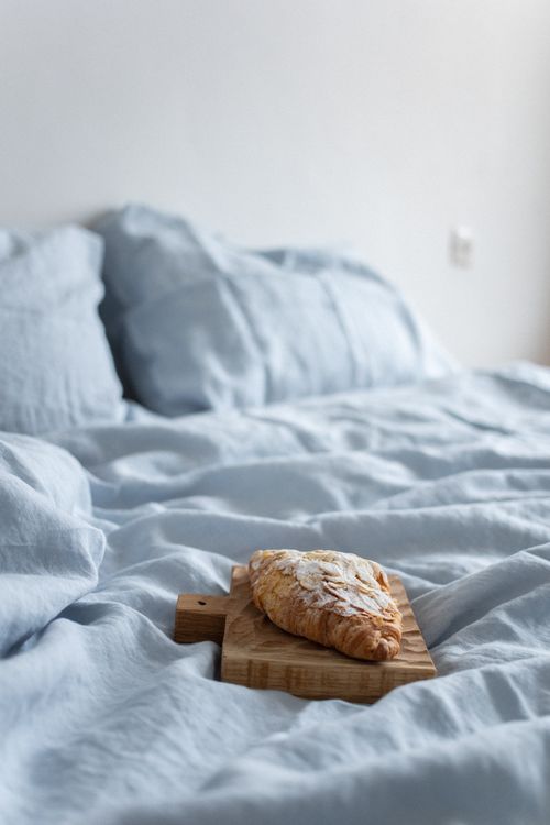 Free A fresh croissant on a wooden board atop a soft, blue bedspread inside a cozy bedroom. Stock Photo