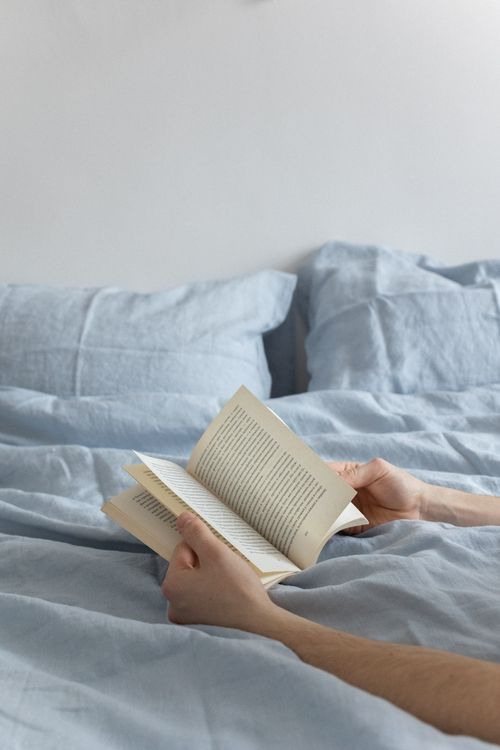 Free Hands holding a book while lounging in a cozy blue-sheeted bed. Stock Photo